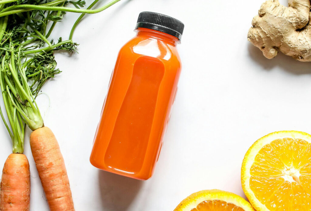 Vegetable juice in a bottle with carrots oranges and ginger surrounding it.